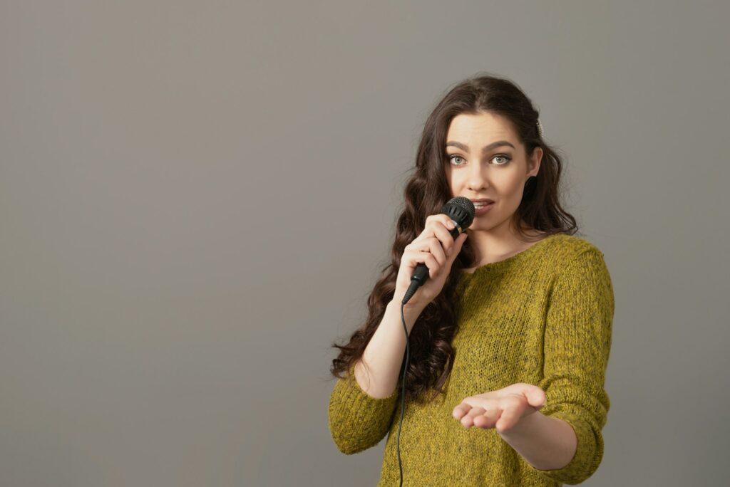 attractive teenager woman speaking with a microphone against gray background, speech presentation